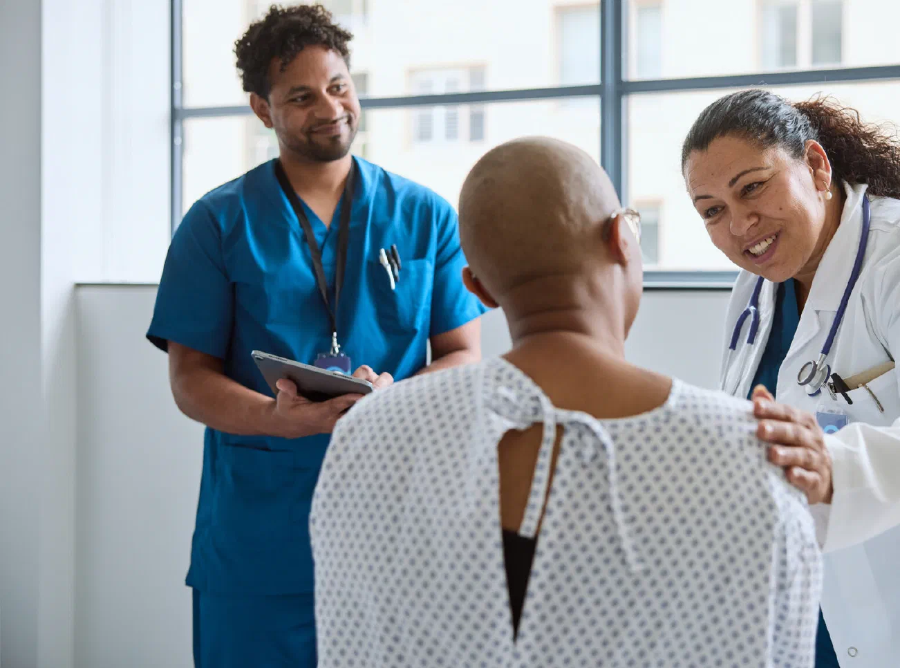 A smiling doctor rests a hand on a patient's shoulder in a clinic while a colleague looks on