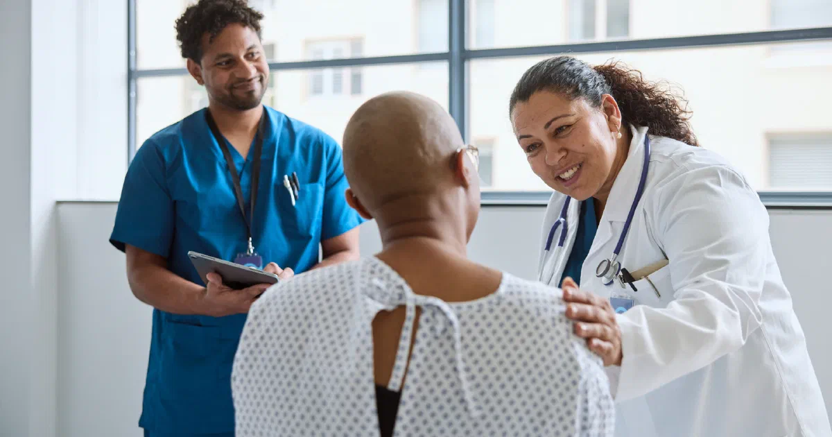 A smiling doctor rests a hand on a patient's shoulder in a clinic while a colleague looks on